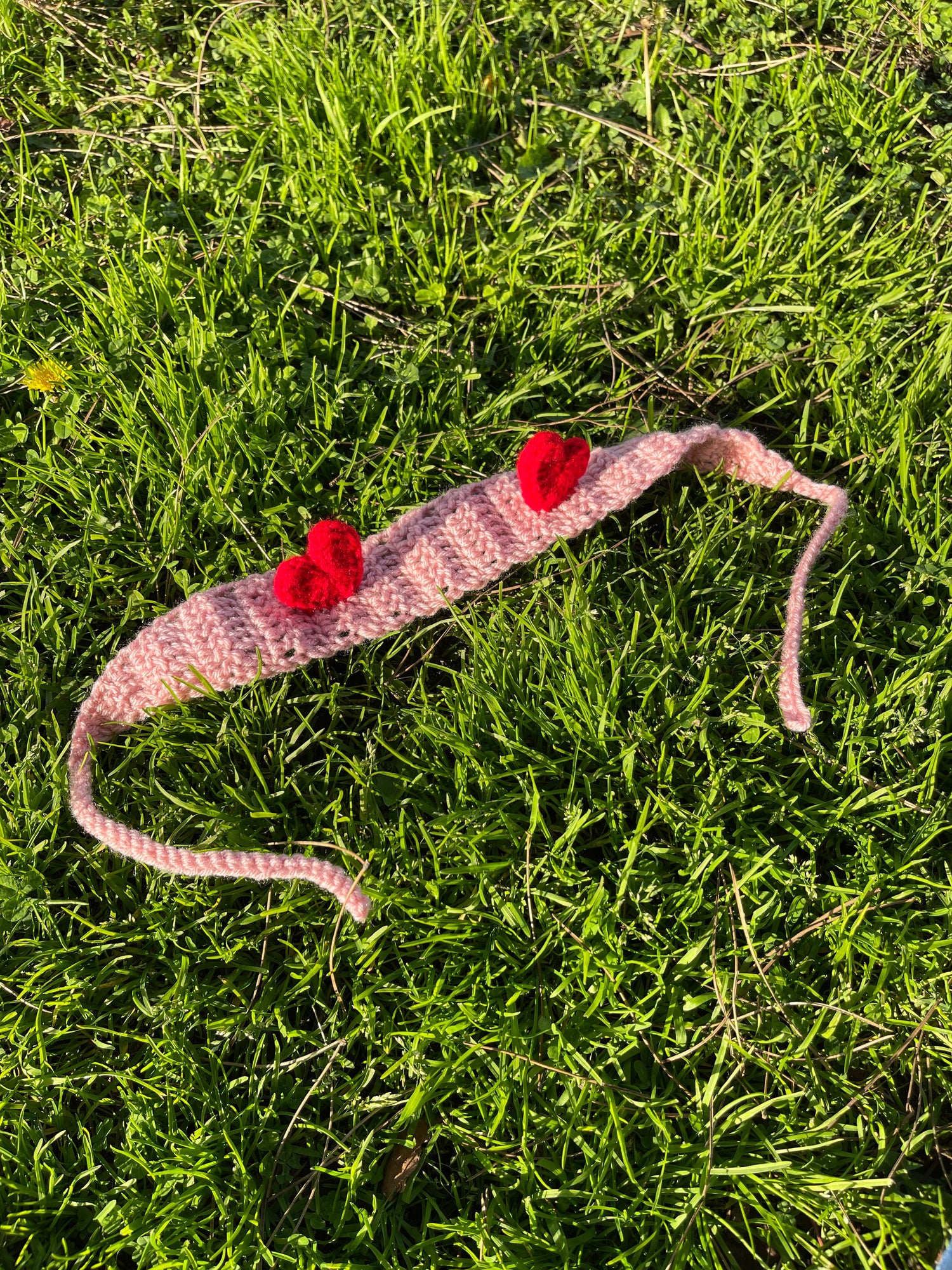 Pink crocheted headband with two red heart decorations placed on green grass.